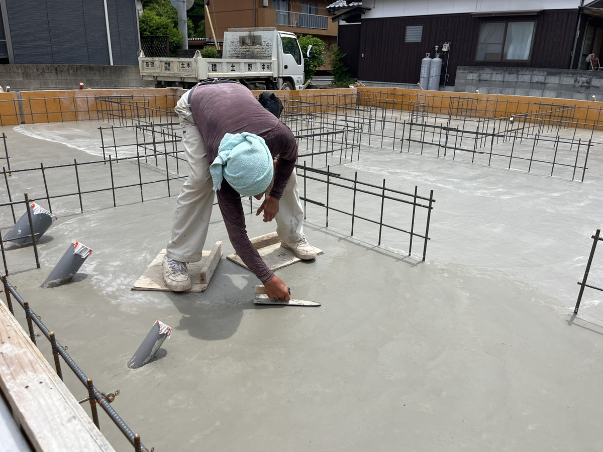 基礎工事の職人が足場板を渡してコンクリートの上に乗り、手作業で丁寧にコテ押さえをして表面を滑らかに均している様子