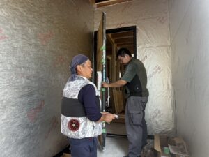 2名の施工スタッフが水平器を使いながら玄関ドアの建て付けを確認している様子。防水透湿シートが貼られた外壁面が見える。